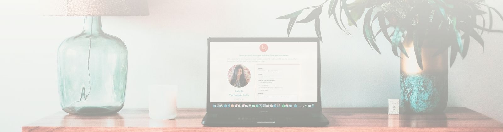 Faded photo showing top of wooden desk with a lamp, vase with leafy plant, and laptop showing The Designist Studio website on screen